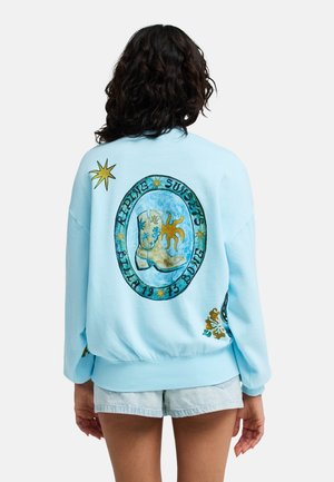 Femme portant un sweat-shirt bleu clair avec un graphisme de botte de cowboy, soleil et le texte "Ripping Sunsets Bllan 1973 Bojong" dans le dos.