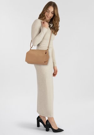Bolso de hombro de piel marrón claro con un acabado texturizado, que cuenta con un cierre de cremallera y una correa ajustable, situado junto a un modelo que lleva un vestido beige.