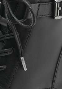 Botín negro de cuero con un acabado suave, puntera rectangular, cordones prominentes y un acento plateado cerca de la parte superior.