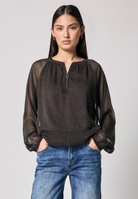 Schwarze transparente Bluse mit einem subtilen Leopardenmuster, runder Ausschnitt und einem Reißverschlussdetails vorne. Die Bluse hat lange Ärmel mit gerafften Bündchen.