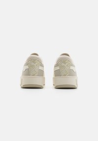 Des baskets beige avec une texture lisse, présentant des accents à motifs sur le talon, deux rayures blanches et un design bas.