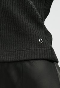 Sweater preto em malha canelada com bainha cosida e um pequeno logótipo prateado "G" no canto inferior esquerdo. Tecido preto suave abaixo.