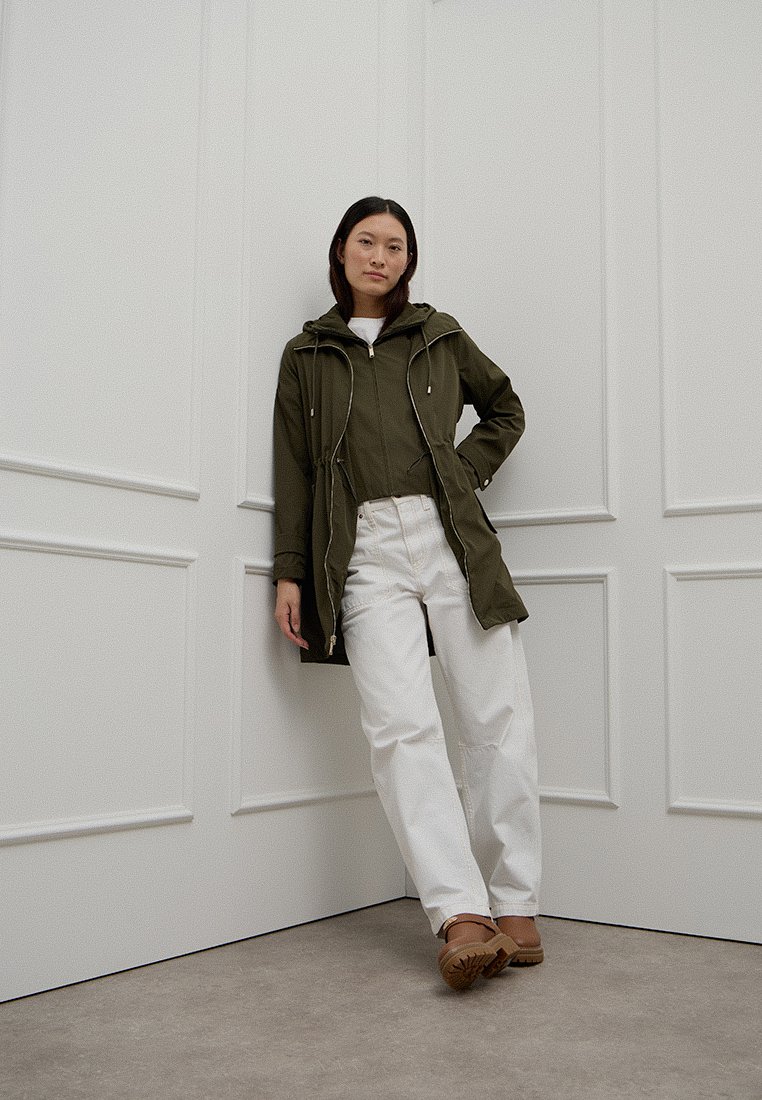 Kobieta oparta o białą panelem ścianę, ubrana w oliwkową kurtkę, białe spodnie i brązowe sandały, stojąca na szarym podłodze.