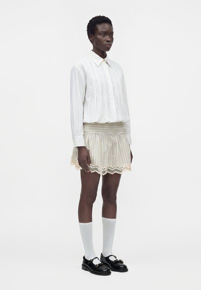 Femme portant une chemise blanche à rayures à manches longues, une jupe beige rayée avec bordure en dentelle, des chaussettes blanches montantes et des mocassins noirs, debout, face à l'avant.