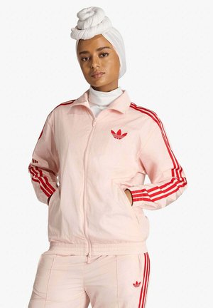 Donna con tuta Adidas rosa con strisce rosse e fascia bianca per capelli annodata in alto, mani nelle tasche della giacca.