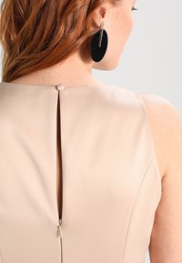 Beige ärmlös klänning med en nyckelhålsöppning bak, liten knappstängning och en strukturerad yta. Har stora svarta örhängen.
