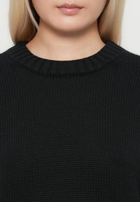 Artimiausias asmens apatinės veido dalies ir kaklo vaizdas, vilkintis juodą raukšlėtą megztinį su apvalia iškirpte.