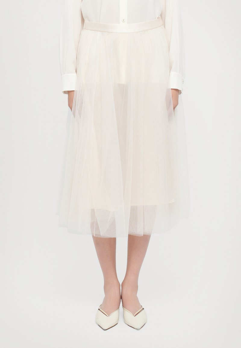 Een knie-lange, crèmekleurige tule rok met een zachte, doorzichtige textuur en een gladde tailleband, gecombineerd met witte, puntige ballerina's.