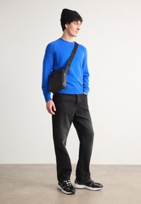Maglione blu a maglia, borsa a tracolla nera, jeans neri e sneakers nere con dettagli bianchi. Il modello indossa un berretto nero.