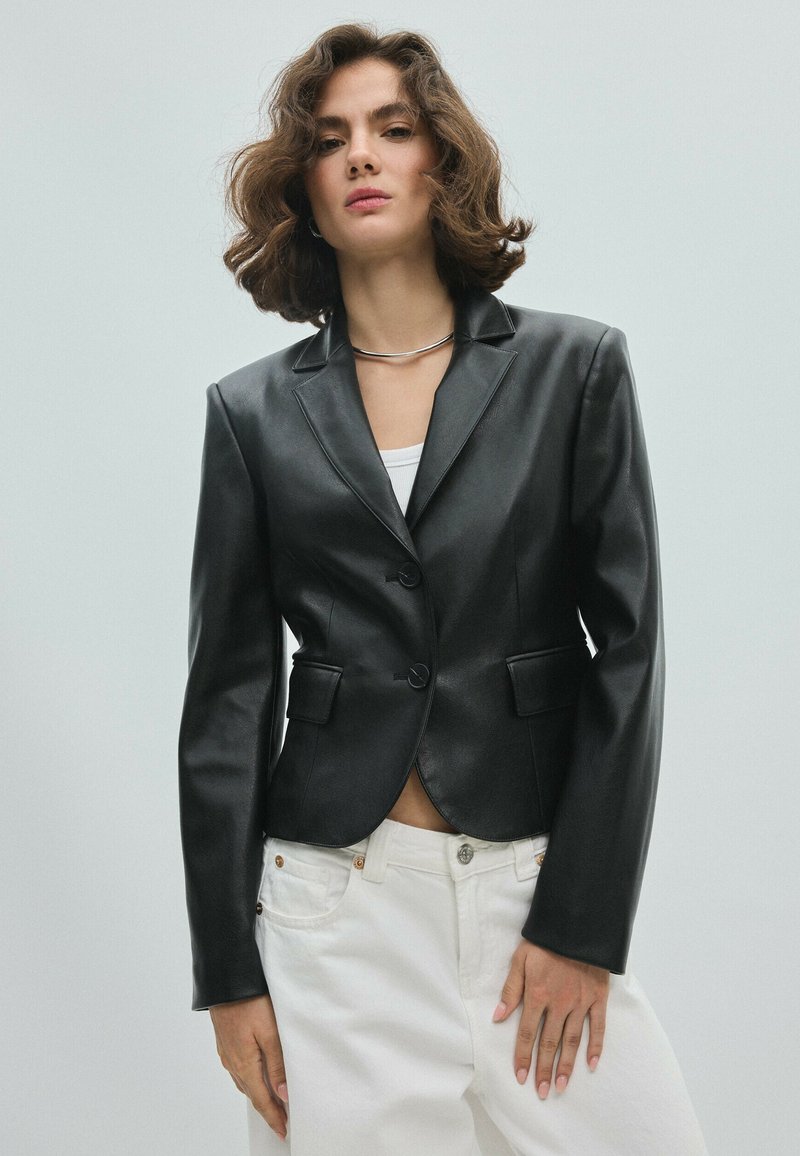 Femme aux cheveux châtain mi-longs porte un blazer en cuir noir par-dessus un haut blanc et un pantalon blanc sur un fond clair uni.