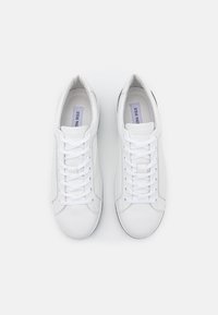 Baskets blanches en cuir avec un design basse, fermeture à lacets plat, texture lisse et un logo discret sur la languette. Flexibilité dans la semelle.