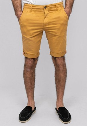 Homme portant un short retroussé jaune moutarde, une chemise blanche, des chaussures noires sans lacets, debout les mains dans les poches sur un fond uni.