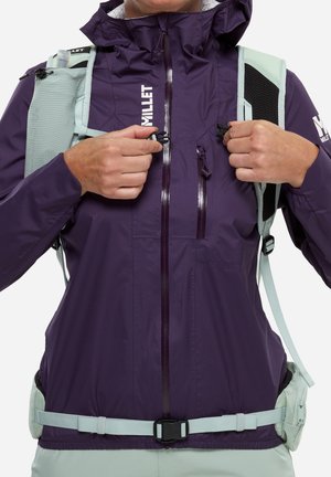 Personne attachant les clips de poitrine sur les bretelles d'un sac à dos gris clair, portant une veste violette Millet avec une fermeture éclair frontale et une poche poitrine.