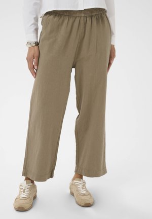 Person iført løstsiddende olivengrønne bukser med elastisk talje og beige sneakers, stående mod en ensfarvet hvid baggrund.