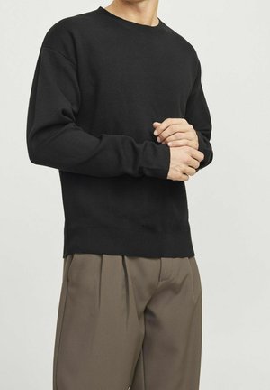 Mann trägt schwarzen Pullover mit langen Ärmeln und locker sitzende braune Hose, mit verschränkten Händen vor einer schlichten hellen Hintergrund.