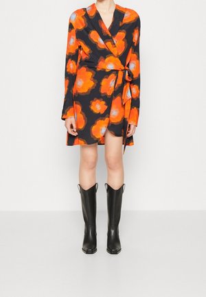 Femme portant une robe portefeuille noire avec de grands motifs floraux orange et gris, associée à des bottes noires montantes sur fond blanc.