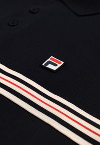 Tissu texturé bleu marine foncé avec deux boutons noirs, détail rayé blanc et rouge, et un petit patch logo rouge et bleu marine.
