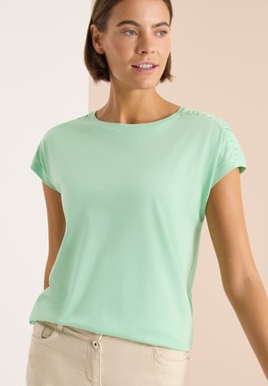 Kvinde iført lysegrøn kortærmet top med rynkede detaljer på skuldrene, kombineret med beige bukser, kigger mod højre mod en neutral baggrund.