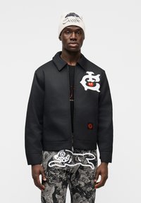 Μαύρο μπουφάν με όρθιο γιακά, διακοσμημένο με λευκά γραφικά στοιχεία και ένα κόκκινο έμβλημα. Συνδυασμένο με παντελόνι γκρι με μοτίβα και πλεκτό σκουφί.