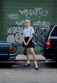 Lyseblå t-skjorte med grafikk, svart denimskjørt, mønstrete veske, hvite sokker og svarte sko med blå detaljer, i urbant miljø.