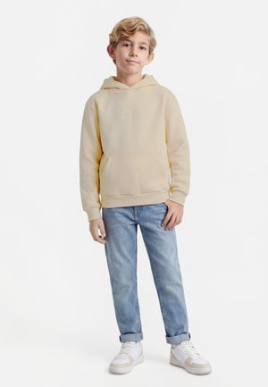 Gaišmatains zēns stāv, valkā bēša krāsas kapuci, gaiši zilas uzrullētas džinsas un baltas sporta kurpes uz vienkārša balta fona.