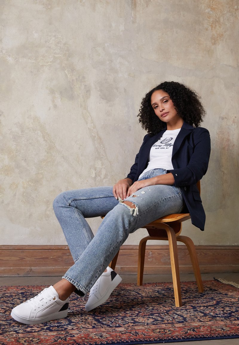 Marineblazer, weißes T-Shirt mit Grafik, ausgewaschene hellblaue Jeans und weiße Sneakers mit schwarzen Akzenten. Modell sitzt auf einem Holzstuhl.