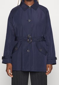 Giacca blu navy con cintura, colletto, chiusura con bottoni, tasche laterali e una texture liscia. Presenta un design strutturato con maniche raglan.