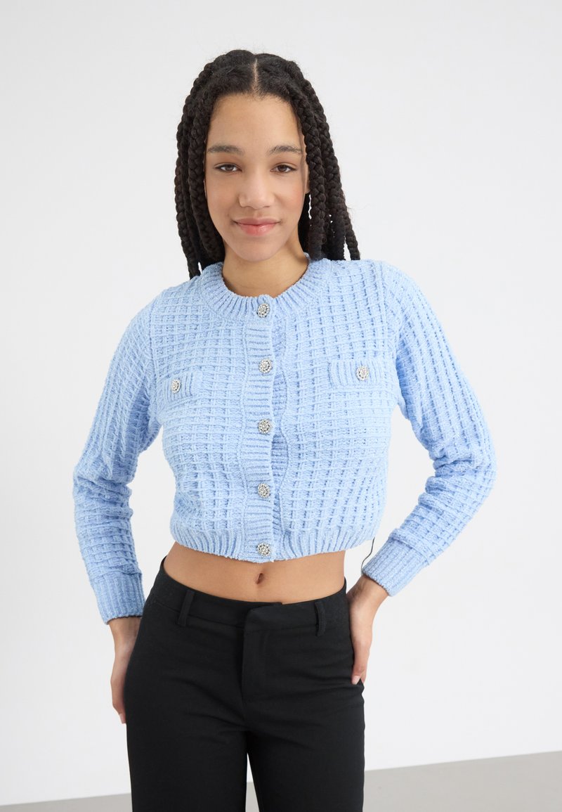 Ljusblå stickad cardigan med en texturerad mönster, kort design, rund halsringning och silverfärgade knappar, kombinerad med svarta byxor.