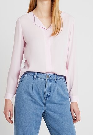 Kobieta ubrana w jasnoróżową, długorękawową bluzkę włożoną w wysokiej talii jasnoniebieskie dżinsy, stojąca na tle jednolitego tła.