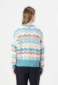 Texturert strikkegenser med bølgemønster i lys blå, krem og oransje, med ribbestrikkede mansjetter og kant. Åpen arbeid design gjennom hele.