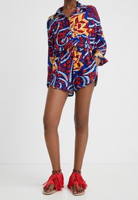 Multicolored kurzer Overall mit einem Kragen, der ein auffälliges Muster in Rot, Blau und Gelb aufweist. Kombiniert mit roten Fransen-Sandalen.