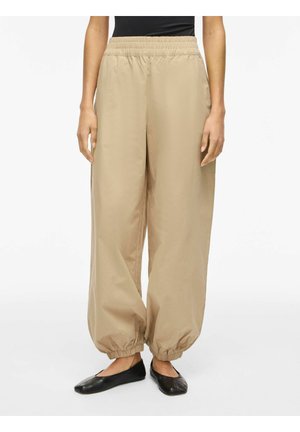 Osoba nosząca luźne beżowe spodnie z elastycznymi ściągaczami przy nogawkach i w pasie, połączone z czarnymi balerinami i czarnym topem.