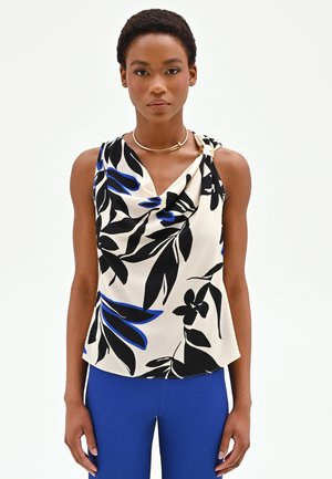 Donna che indossa una canottiera bianca senza maniche con stampa di foglie nere e blu e pantaloni blu, rivolta in avanti su uno sfondo bianco.