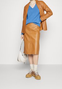 Brauner Lederblazer kombiniert mit einem blauen V-Ausschnitt-Pullover und einem braunen Lederrock. Klobige beige Schuhe und eine weiße Handtasche runden das Outfit ab.