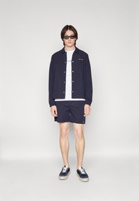 Marineblå skjorte med knapper over en hvid t-shirt, parret med marineblå shorts og marineblå sneakers. Har minimalistisk design og diskret branding.