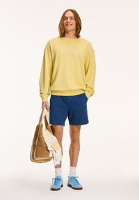 Žltá mikina s embosovaným textom, námornícke šortky, svetlomodré tenisky a tkaná taška s prúžkovaným uterákom. Jednoduchý, neformálny outfit.