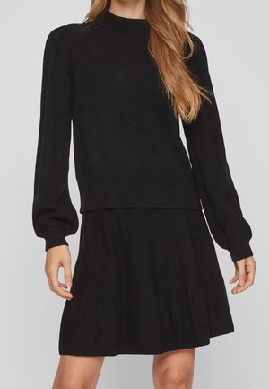 Woman wearing a black long-sleeve knit sweater with a matching black skirt, standing against a plain light gray background.