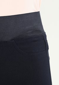 Námořnické modré kalhoty s širokým elastickým černým pasem, boční kapsou a hladkým povrchem, zdůrazňující pružnost látky.