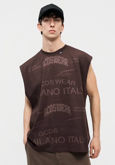 Νεαρός άνδρας που φοράει αμάνικο σκούρο καφέ πουκάμισο με το κείμενο "GCDS WEAR MILANO ITALY" και μαύρο καπέλο, στέκεται με το ένα χέρι στην τσέπη.