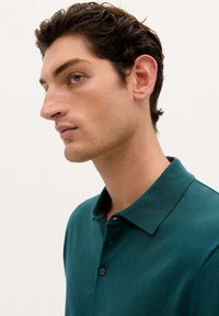 Grøn polo t-shirt lavet af struktureret stof, med krave, tre knapper og en subtil logo-detalje på venstre side.