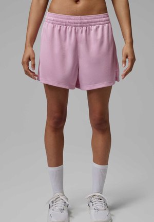 Personne portant un short de sport rose clair, des chaussettes blanches hautes et des baskets blanches, debout devant un fond gris uni.