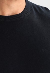 Nahaufnahme einer Person, die einen schwarzen Strickpullover mit Rundhalsausschnitt trägt, auf dessen Brust dezent der Text "ESPR" gestickt ist.