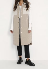 Beige gesteppte Weste mit zwei großen Fronttaschen, Druckknöpfen und einem Kragen, getragen über einem weißen Langarmshirt und schwarzen Hosen.