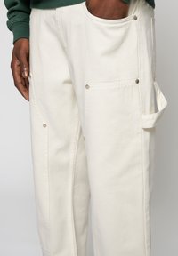 Pantalones de algodón blancos con múltiples bolsillos, broches de botón plateados y un lazo lateral para sujeción. Textura suave y corte relajado.