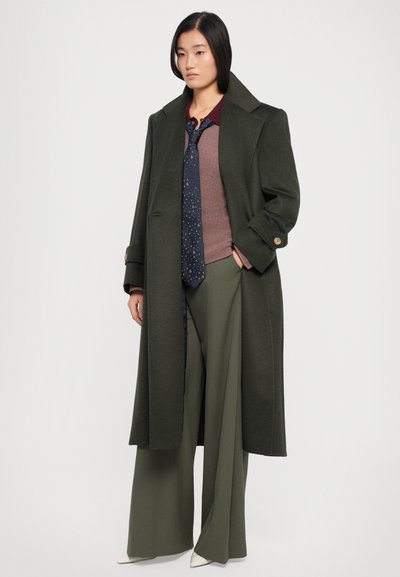 Long manteau en laine vert olive avec un grand col et des poignets boutonnés, superposé sur un haut marron à côtes et un pantalon large olive, accompagné d'une cravate à motifs.