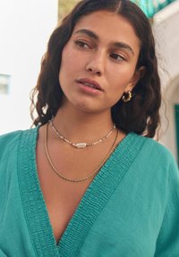 Vêtement en teal texturé avec un col en V, orné de colliers superposés : une chaîne en or, une chaîne de perles avec une pierre ovale, et une chaîne dorée mince torsadée.