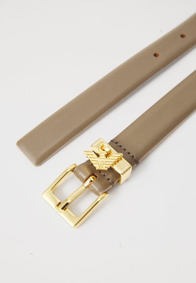 Ceinture en cuir brun clair avec boucle en or et emblème représentant un design ailé, posée à plat sur une surface blanche.