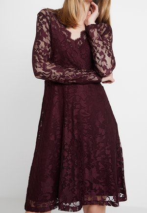 Vrouw in een bordeauxrode kanten jurk met lange mouwen en een lengte tot de knieën, met bloemmotieven, poseert terwijl ze met de ene hand haar gezicht aanraakt en de andere hand gekruist houdt.