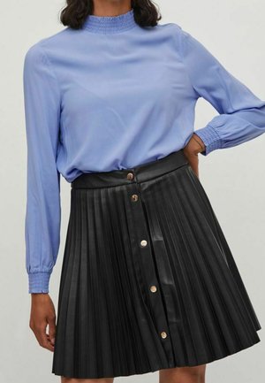 Vrouw draagt een lichtblauw shirt met lange mouwen, voorzien van een geplooide kraag en manchetten, gecombineerd met een zwarte geplooide rok met gouden knopen aan de voorkant.