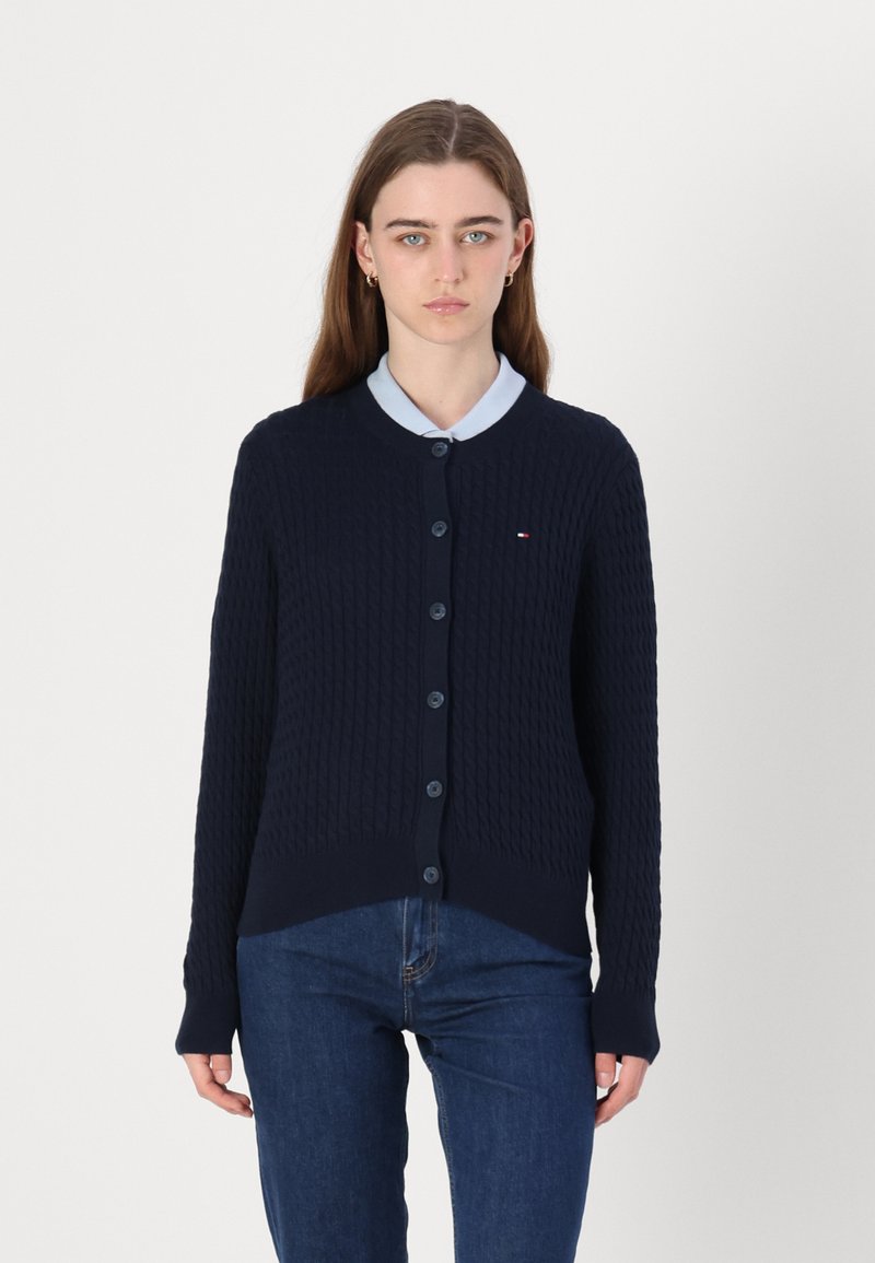 Ναυτικό πλέκτο cardigan με στρογγυλή λαιμόκοψη, κλείσιμο με κουμπί, ριμπ τελείωμα και υφασμάτινο σχέδιο. Φορεμένο πάνω από ένα ανοικτό μπλε πουκάμισο.